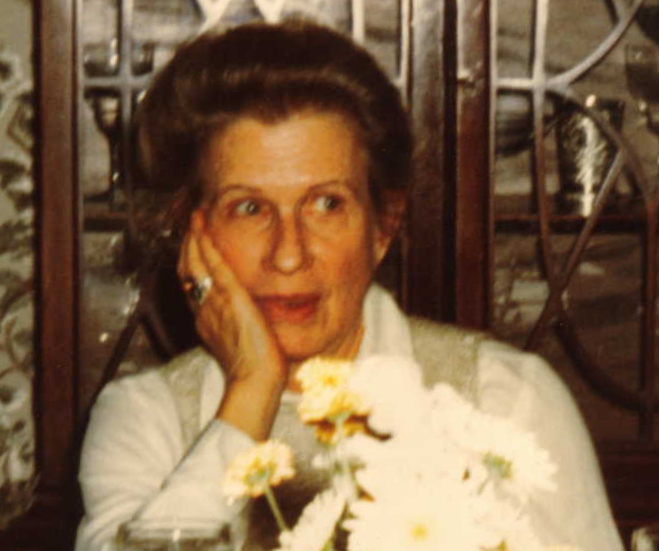 Mildred rests her head against her right hand, elbow on a table we cannot see. A few dasies in the foreground slightly obscure her.