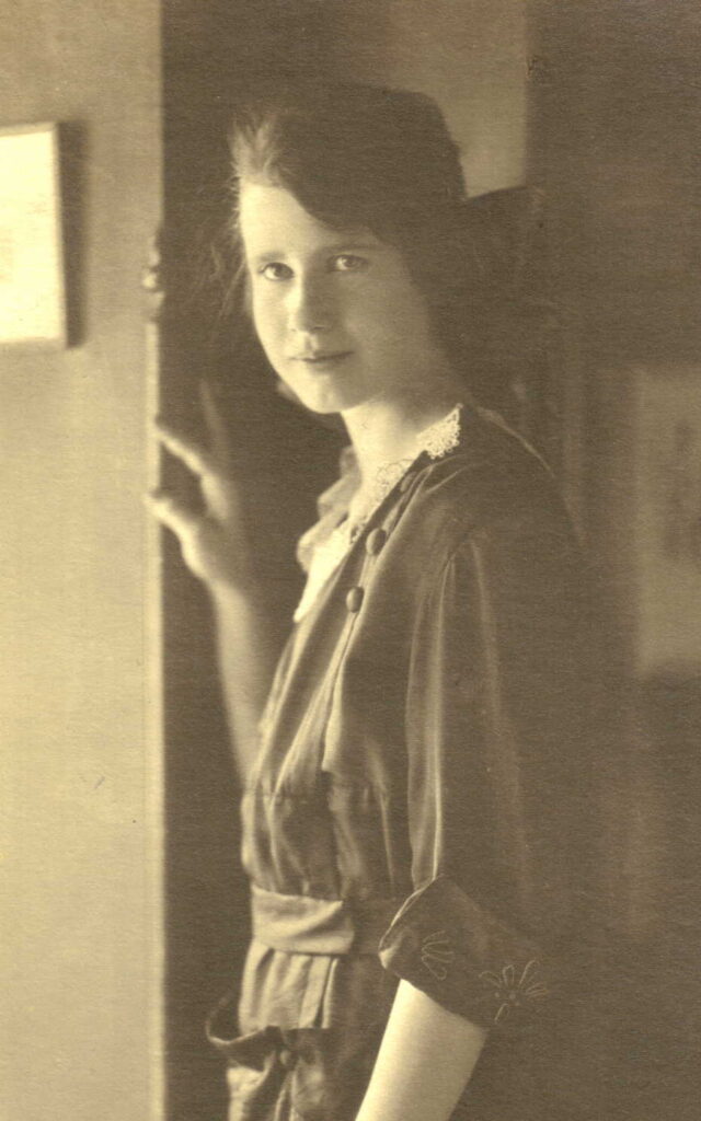 A young mildred stands in a doorway, left hand resting on the frame smiling gently at the camera.