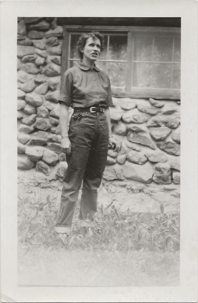 Black and white photograph of a woman in rolled up jeans and a short-sleeve shirt standing in front of a stone cabin.